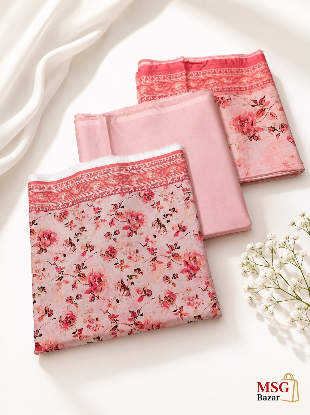 Three folded pieces of pink fabric with floral prints and patterned borders, arranged on a white surface with draped white fabric and small white flowers. A 'MSG Bazar' logo appears in the bottom right corner.