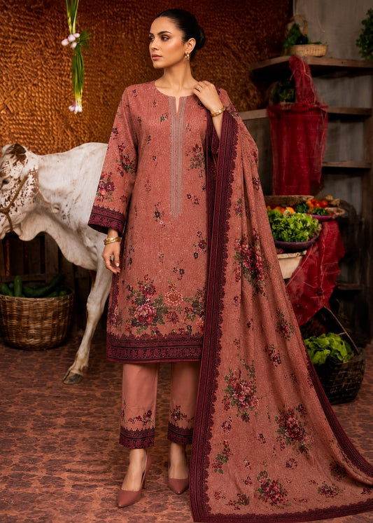 A woman models a three-piece salwar kameez suit in dusty rose with floral prints and dark borders, paired with matching trousers and a long dupatta, set against a rustic indoor backdrop with a cow and decorative elements.