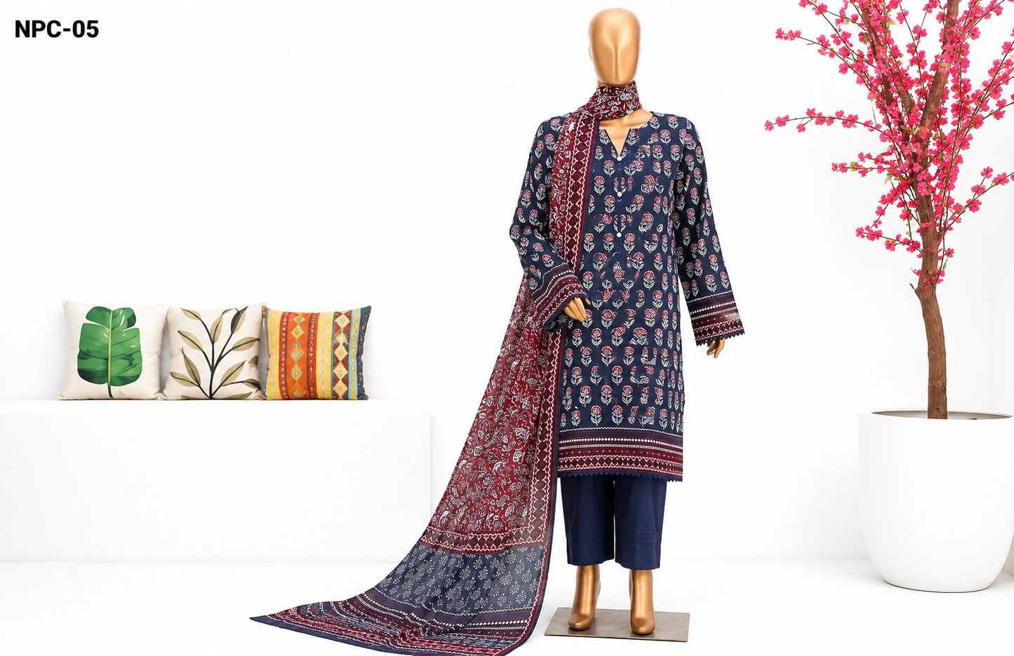 A three-piece Indian ethnic suit set displayed on a mannequin, featuring a navy blue kurta with red and gold floral prints, matching navy pants, and a coordinating dupatta. The set is styled in a clean studio setting with decorative pillows and a potted pink tree.