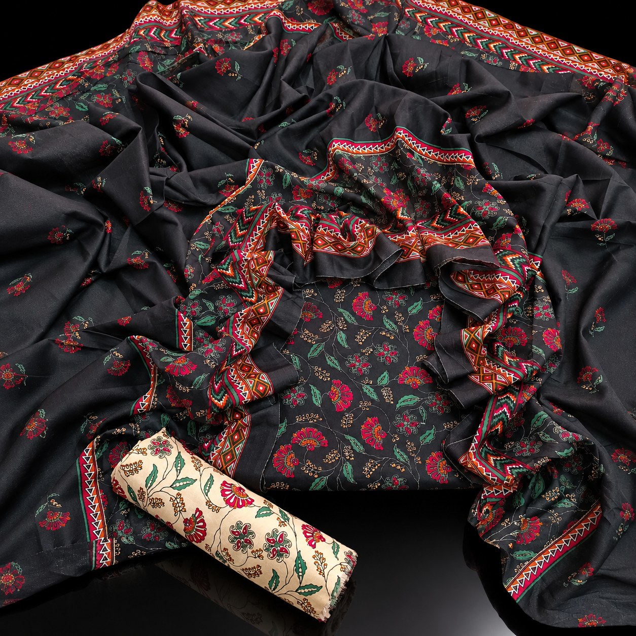 A black floral printed fabric set with red and green floral motifs, decorated with red and gold patterned borders, shown draped on a black surface with a rolled beige fabric piece featuring matching floral print.