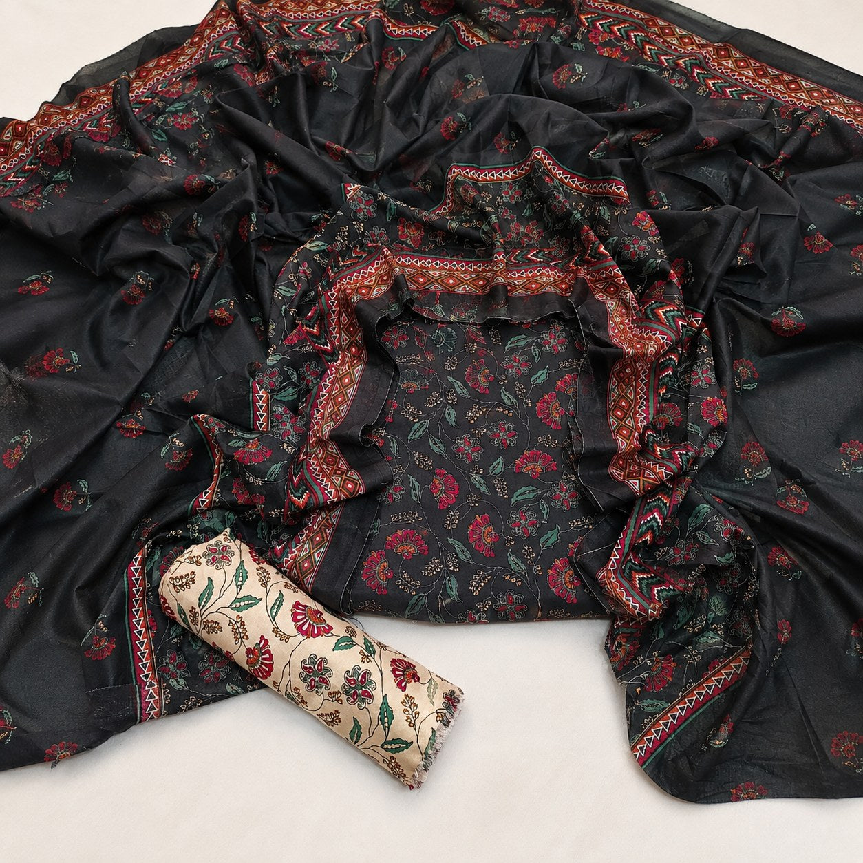 Black cotton fabric set with red and green floral block prints, featuring a matching printed dupatta with a red patterned border and a rolled beige fabric piece with floral motifs.
