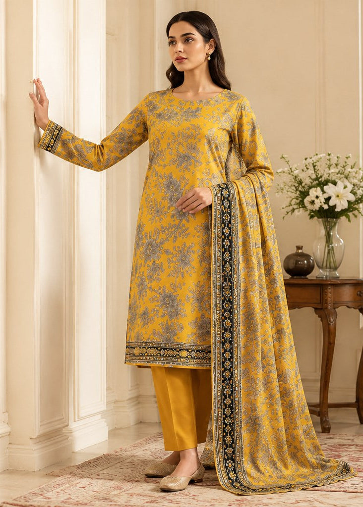 A woman models a traditional Indian salwar kameez suit in a warm beige interior setting. The outfit consists of a mustard yellow tunic with a blue floral print and a matching yellow trouser pant, accented by a yellow dupatta with a black and gold border.