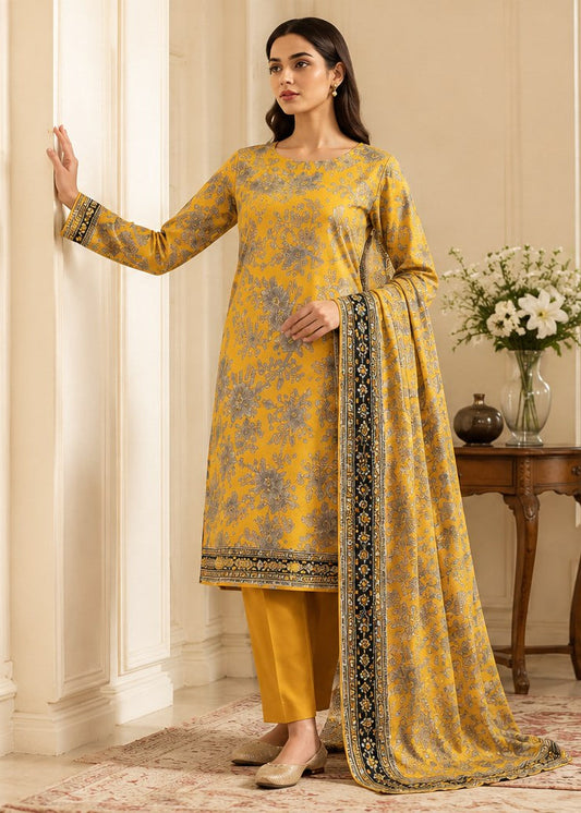 A woman models a traditional Indian salwar kameez suit in a warm beige interior setting. The outfit consists of a mustard yellow tunic with a blue floral print and a matching yellow trouser pant, accented by a yellow dupatta with a black and gold border.