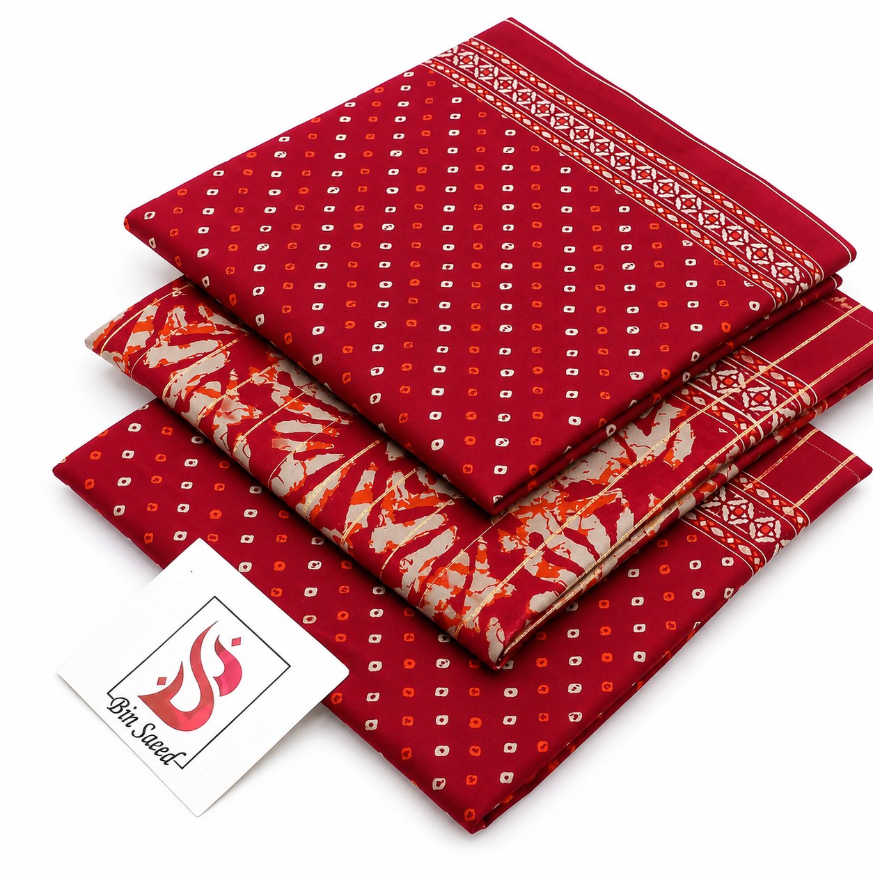 Three folded red patterned fabric pieces stacked on a white background, with a small white brand tag reading 'Lotus Saffron' in the lower left. The fabrics show small white and orange motifs and decorative border patterns.