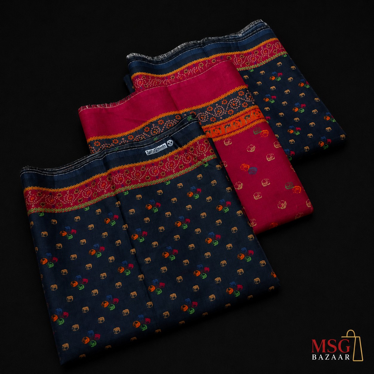 Three folded navy blue sarees with colorful floral patterns and bright pink borders, arranged diagonally on a black background. A 'MSG BAZAAR' logo is visible in the bottom right corner.