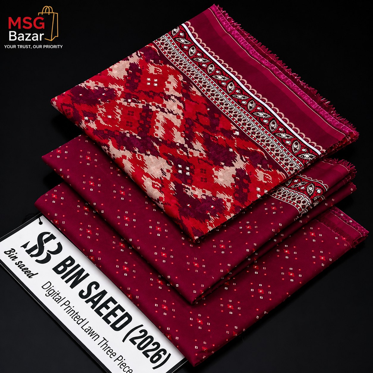 Three folded pieces of printed lawn fabric from MSG Bazar displayed on a black background, featuring a red and cream geometric patterned piece on top, a solid deep red piece in the middle, and a dark red dotted piece on the bottom. A white promotional banner in the corner reads '3 BIN SALEED (2026) Digital Printed Lawn Three Piece'.