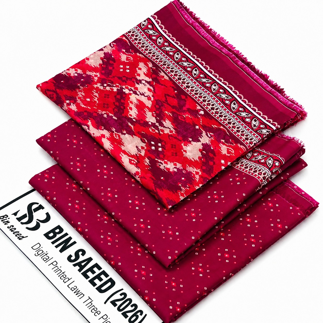 Three folded fabric pieces in a stacked arrangement on a white background, with a printed paper label at the bottom left reading '53 BIN SAEED Digital Printed Lawn Three Piece'. The top fabric has a red, pink, and cream geometric pattern with a decorative border; the middle and bottom fabrics are solid deep pink with small white dot prints.