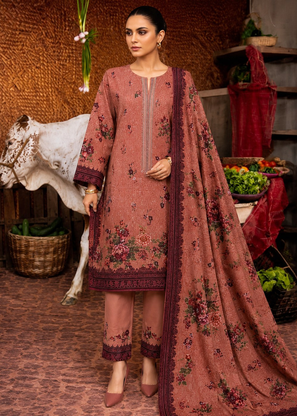 A woman models a traditional three-piece salwar kameez suit in dusty rose pink with floral embroidery, featuring a long-sleeve kurta, matching trousers, and a coordinating dupatta, set against a rustic indoor backdrop with a cow and decorative elements.