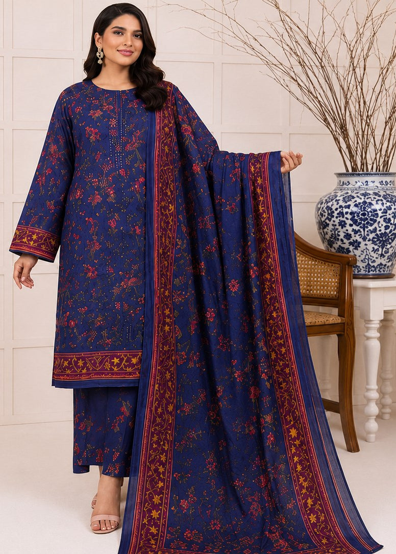 A woman models a navy blue floral printed salwar kameez suit with a matching dupatta, featuring red and gold border details, in a styled indoor setting with a tiled wall, wooden chair, and decorative vase.