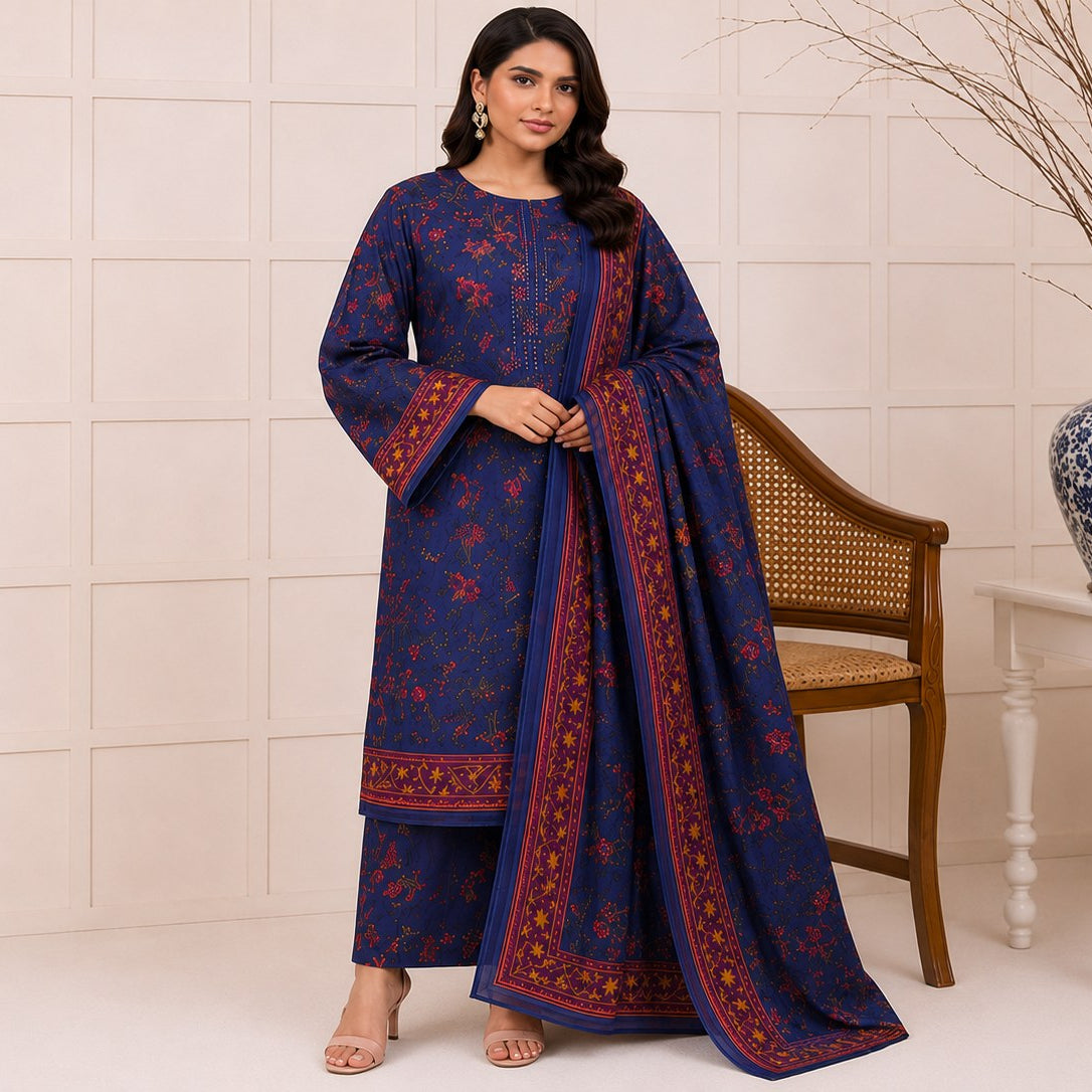A woman models a deep blue three-piece ethnic suit with red floral patterns and border details, standing in a studio setting with a chair and a decorative blue-and-white vase in the background.