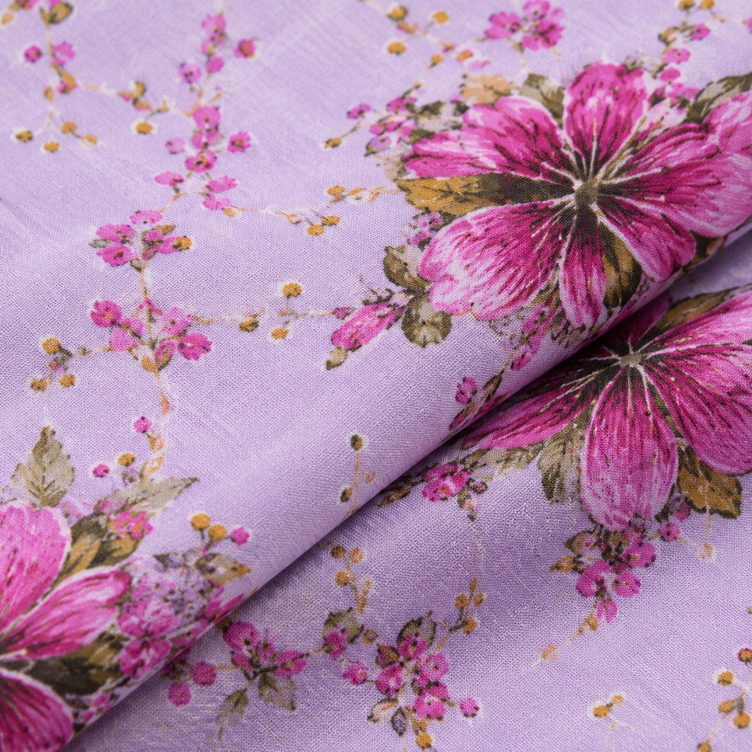 A close-up of lavender purple fabric with a pink floral print, featuring large magenta flowers and small pink blossoms with green and gold accents, draped with a fold in the center.