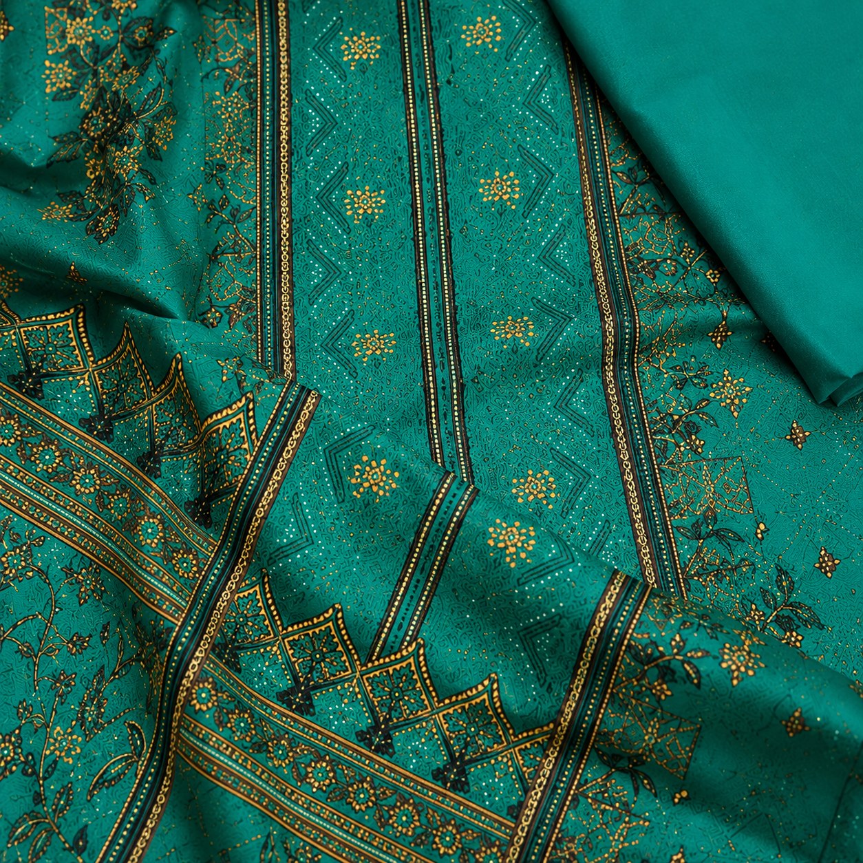 A close-up of a teal green fabric with intricate gold and black geometric embroidery and floral motifs, with a matching solid teal fabric folded in the upper right corner.