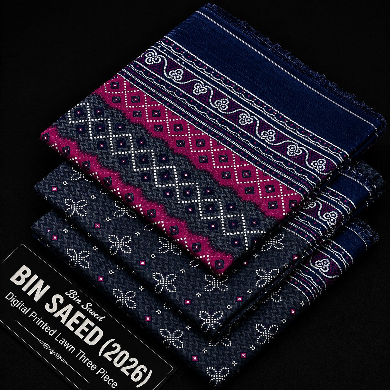 Three folded patterned fabric pieces stacked on a black background, with a black text label reading 'BIN SAEED (2026)' and 'Blue Saeed Digital Printed Lawn Three Piece.' The fabrics show dark navy and charcoal woven-style patterns with bright pink/magenta bands and white decorative motifs.