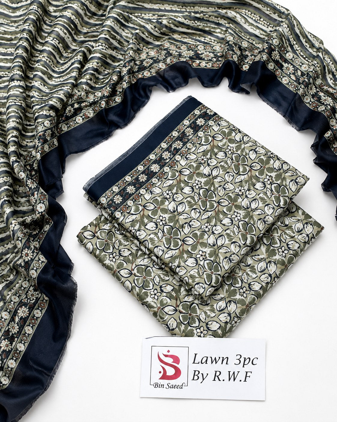 A folded and draped three-piece cotton fabric set with a dark navy blue border and a green, white, and gray floral block print. A white label at the bottom reads 'Lawn 3pc By R.