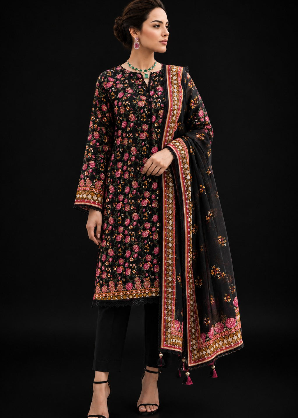 A woman modeling a black three-piece ethnic suit with pink and gold floral embroidery, featuring a long kurta, black trousers, and a matching dupatta, set against a solid black background.