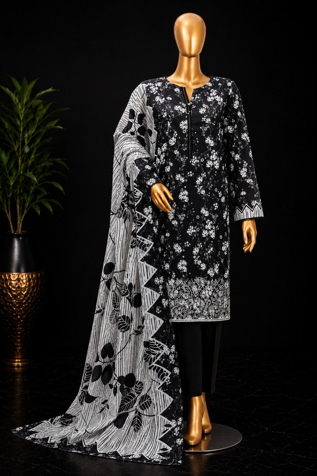 A black and white printed three-piece women's suit displayed on a gold mannequin against a black background, with a potted plant on the left. The set includes a black kurta with white floral print, black trousers, and a matching dupatta with black and white abstract patterns.