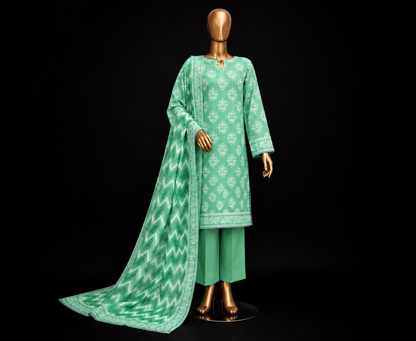 A three-piece green ethnic suit displayed on a gold mannequin against a black background. The set includes a long-sleeved kurta with white floral embroidery, matching green trousers, and a long dupatta with a white chevron pattern and border.